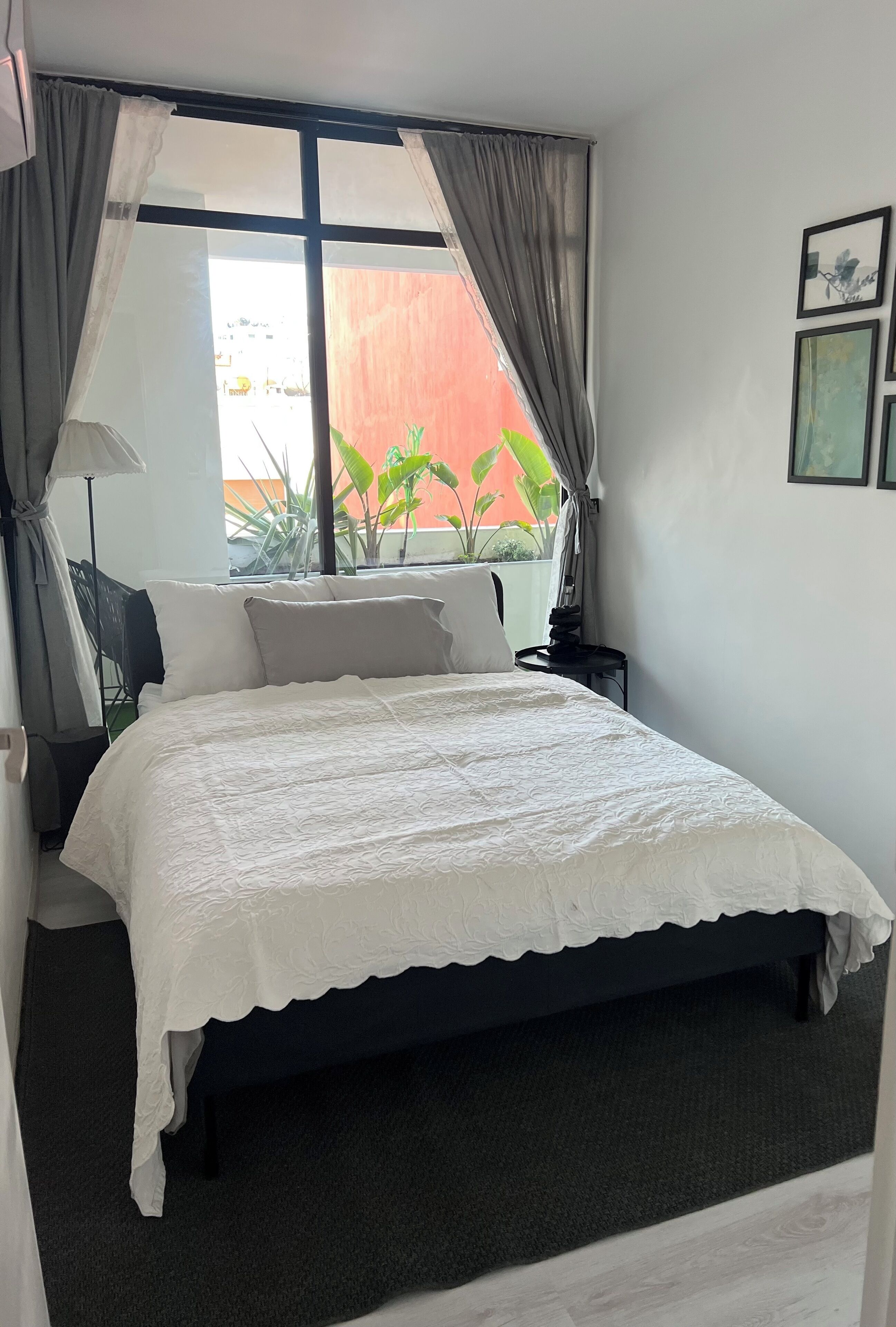 Un dormitorio con cama, cortinas, una mesita de noche, lámpara y ventana con vistas a plantas y un edificio.