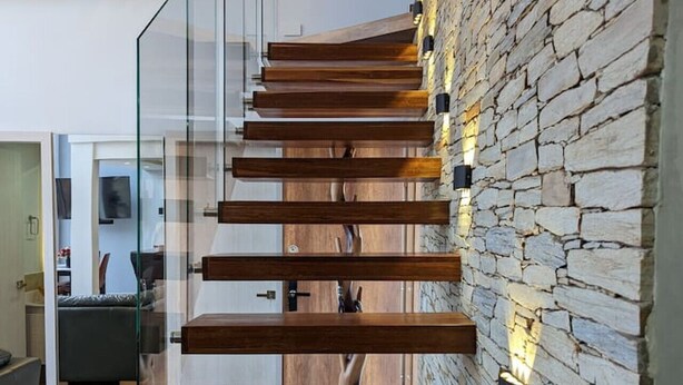 A modern staircase with glass railings and wooden steps, leading to a room with a stone wall and a flat-screen TV. - Exclusive Luxury In Heart of Medellin. (Medellín)