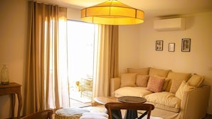 Un salon avec un canapé beige, une table ronde et une chaise. On y trouve une grande fenêtre avec des rideaux, un climatiseur fixé au mur et deux tableaux encadrés.