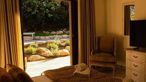 A room with a view of a garden, a wooden bench, a chair, a television, and a bed with a rolled-up towel.