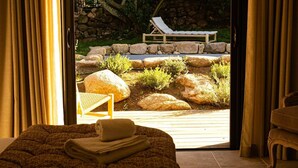 A room with a bed, a window with curtains, and a view of an outdoor area with a lounge chair and rocks.