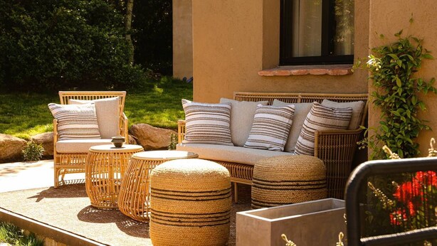 An outdoor seating area with wicker furniture and cushions, a stone planter, and a window with a view of greenery.