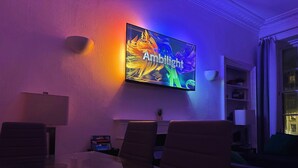A modern living room with a flat-screen TV mounted on the wall, a glass-top dining table, leather chairs, and a bookshelf.