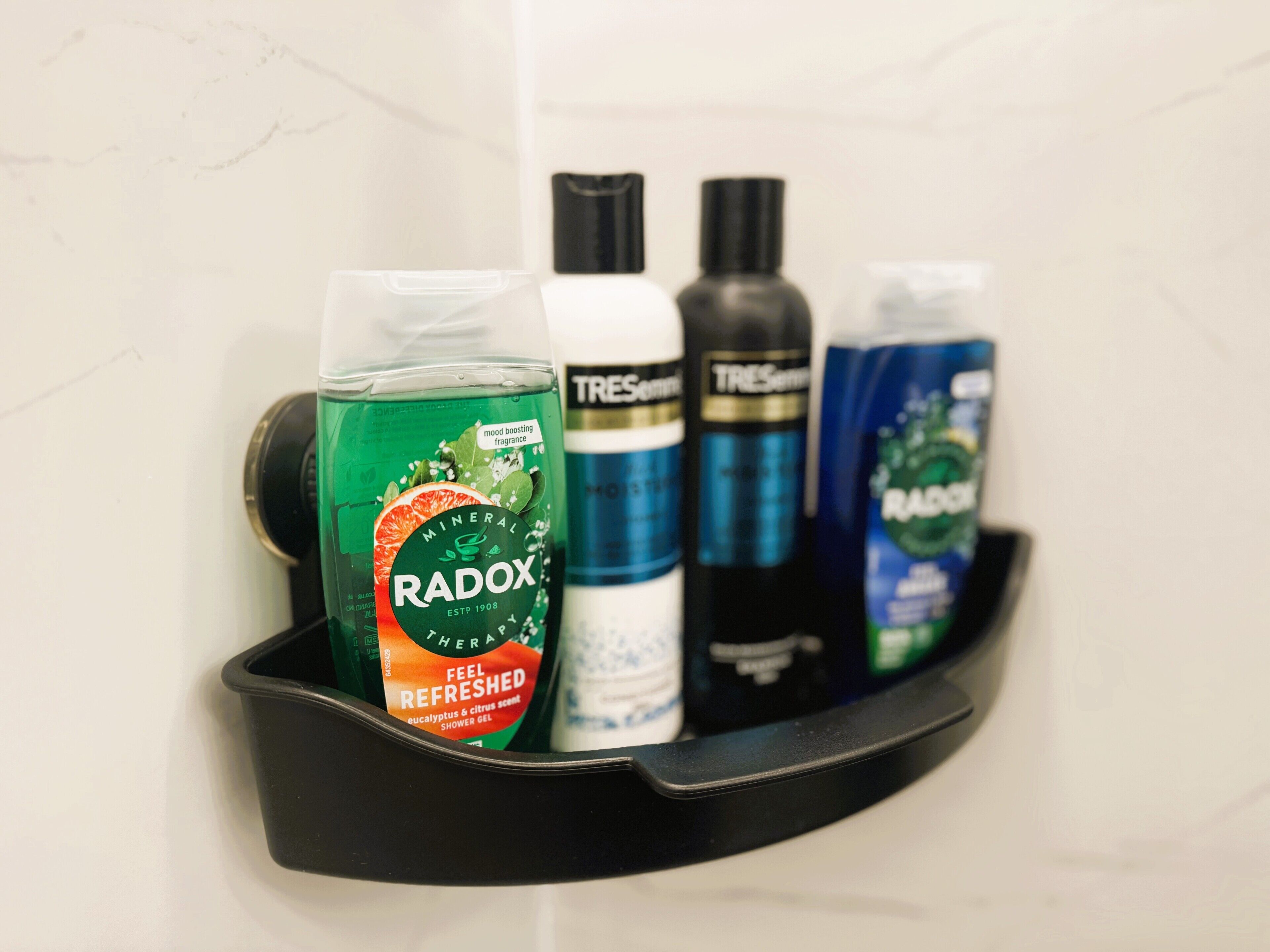Estante de baño con varios artículos de aseo, incluyendo una botella verde etiquetada 'Radox', una blanca, una negra y una azul.