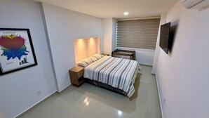 Ein Schlafzimmer mit Bett, Nachttischen, einem an der Wand montierten Fernseher und einem Fenster mit Jalousien.