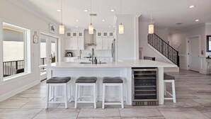 Una cocina amplia con una isla central, muebles blancos, iluminación colgante y un refrigerador de acero inoxidable.