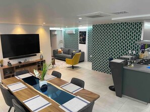 A modern living room with a large flat-screen TV, a dining area with a long table, and a kitchen area with a counter and appliances. - Modern Luxury Fortress with 418 Mbps Fiber Wifi and Secure Garage (Medellín)