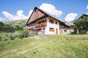 A traditional wooden house with a balcony, surrounded by a fenced yard and a hilly landscape.