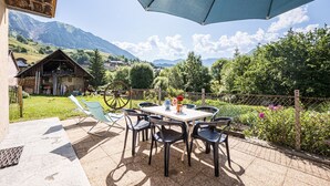 An outdoor patio with a table and chairs, a blue umbrella, and a view of mountains and greenery.