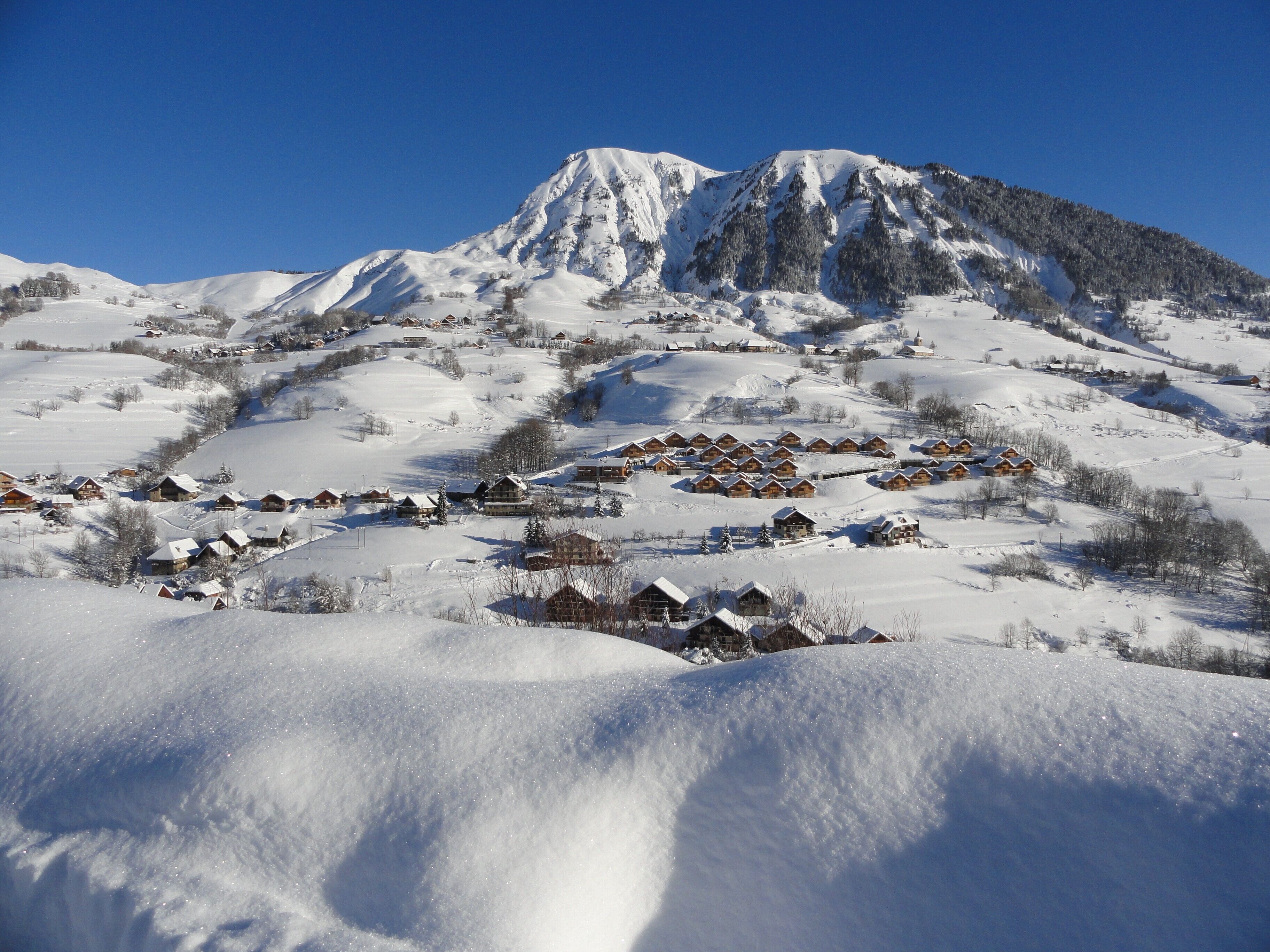 Une montagne enneigée sous un ciel dégagé, un village aux maisons en bois et un sommet imposant.
