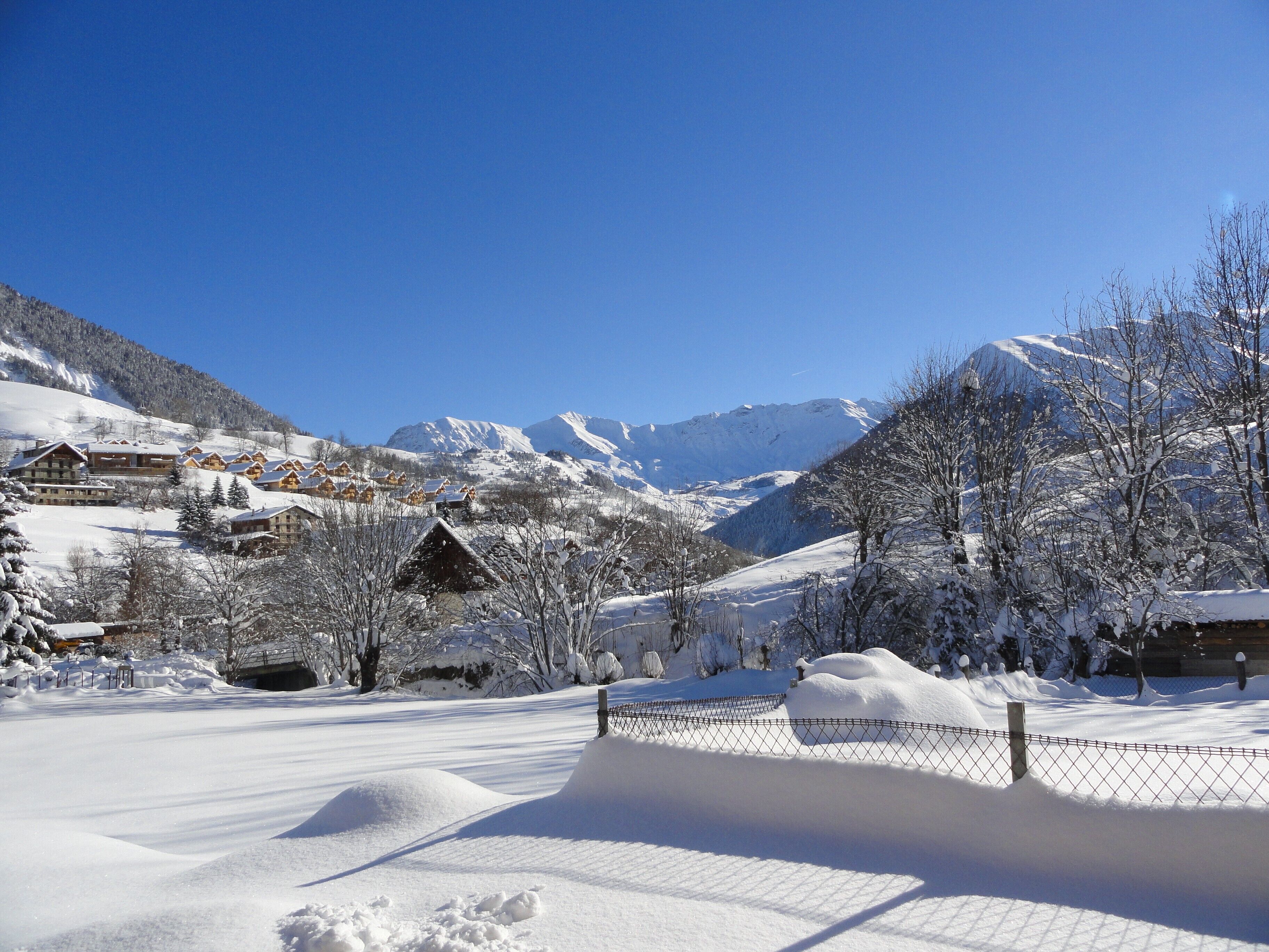 Un pueblo en la montaña cubierto de nieve, con edificios de estilo chalet, una zona cercada y un cielo azul despejado.