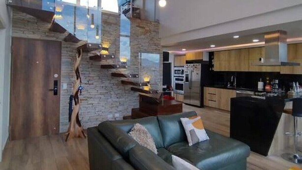 Amplia sala de estar con diseño moderno, que incluye un sofá de cuero, una escalera con pared de piedra y una cocina al fondo.