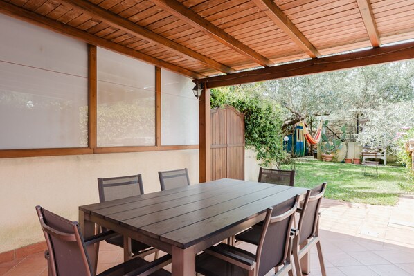 An outdoor patio with a wooden table and chairs, a covered area with a wooden ceiling, and a view of a garden with plants and a fence.