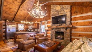 A log cabin interior with a stone fireplace, wooden ceiling, and a kitchen area.