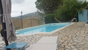 Piscina com piso de pedra, área coberta com uma cadeira e vista para colinas.