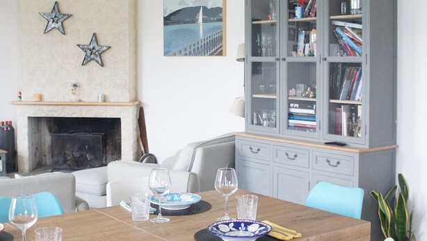 Uma sala de jantar com mesa de madeira, cadeiras azuis e brancas, lareira, estante de livros e um candelabro.
