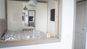 Quarto com cama, mesa de cabeceira, espelho e janela com vista para um corredor e uma porta.