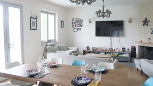 Uma sala de estar com uma mesa de jantar posta, um sofá, uma televisão fixada na parede e uma lareira.