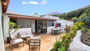 Um pátio com um sofá, cadeiras e uma mesa de jantar, rodeado por plantas e vista para uma montanha.