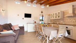 Uma sala de estar aconchegante com um sofá, uma mesa de jantar com cadeiras, uma cozinha com armários e eletrodomésticos, e uma TV fixada na parede.