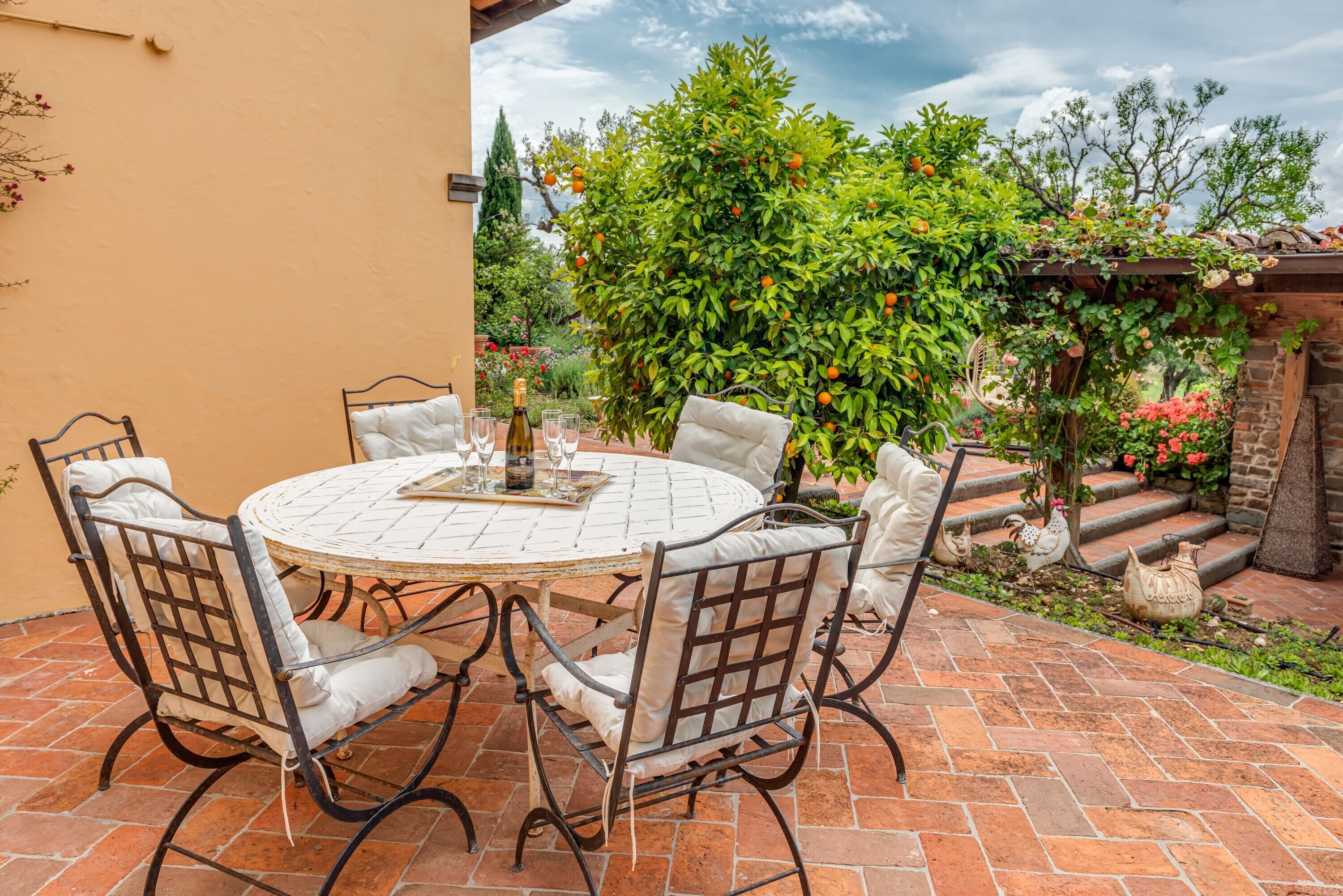 A round table with four chairs, a bottle of wine, and glasses on a patio with a brick floor.