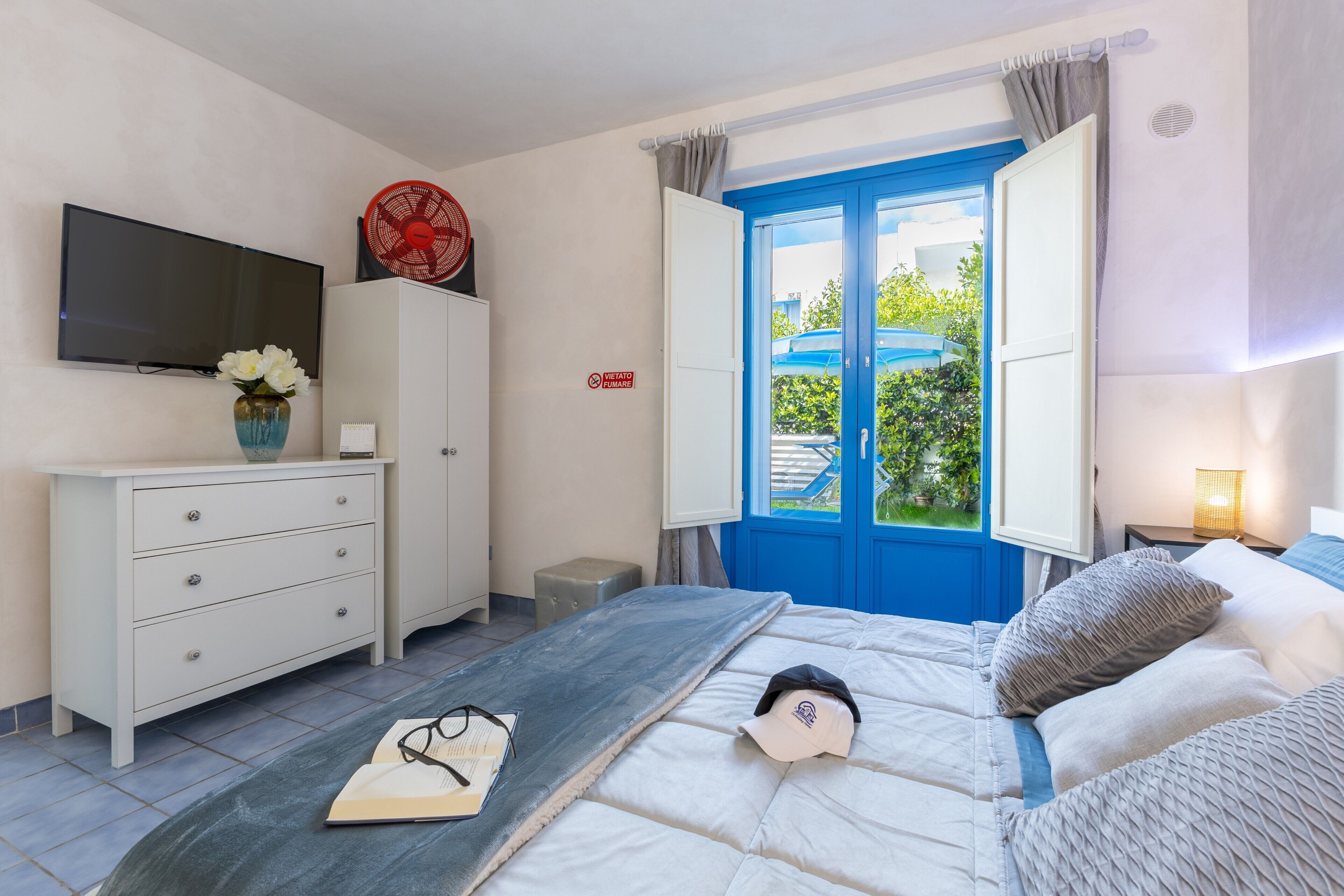 A bedroom with a bed, a TV, a dresser, a small table with a vase of flowers, and a window with blue shutters.