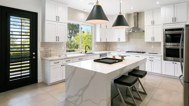 Una cocina moderna con armarios blancos, una isla grande, lámparas colgantes negras y electrodomésticos de acero inoxidable.