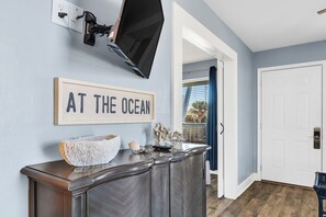 A room with a wooden console table, a flat-screen TV mounted on the wall, and a framed sign that reads "AT THE OCEAN".