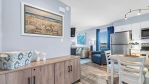 Sala de estar moderna com uma pintura grande, área de jantar com cadeiras brancas e cozinha com eletrodomésticos de aço inoxidável.