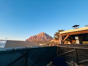 Un paysage montagneux sous un ciel dégagé, un bâtiment doté d’une terrasse en bois et une vue sur un pont au loin.