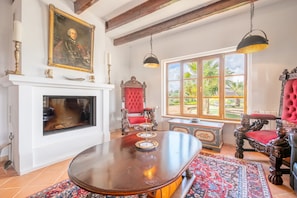 A room with a fireplace, ornate chairs, a large table, and a window with a view of greenery.