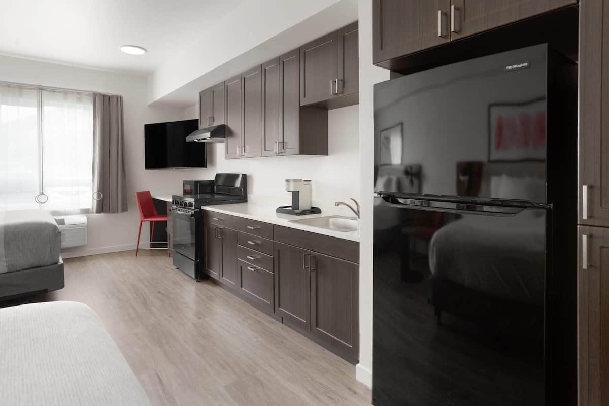 A modern hotel room with a kitchenette, a bed, a red chair, and a large window with curtains.