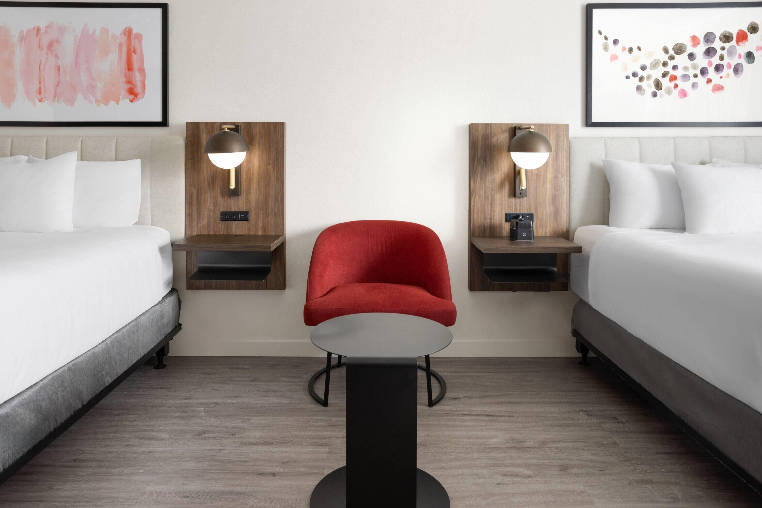A hotel room with two beds, a red chair, a small table, and two wall-mounted lamps.
