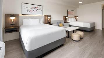 Une chambre d’hôtel comprenant un grand lit, un lit plus petit, un canapé, une table basse et deux poufs. Des lampes murales et des œuvres d’art sont accrochées aux murs.