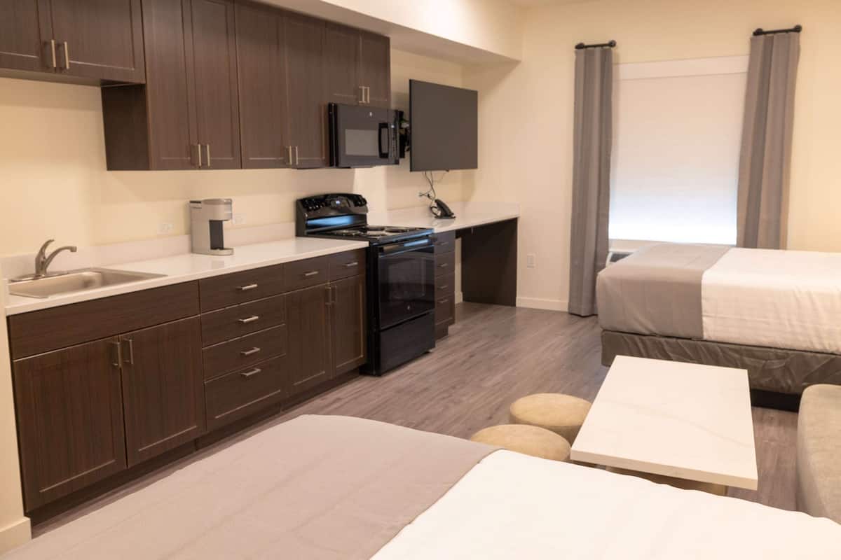 A compact hotel room with a kitchenette, a bed, a bedside table, and a window with curtains.