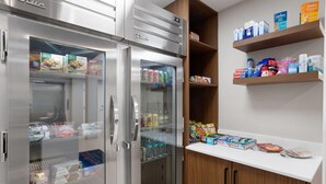 Una kitchenette con un refrigerador de acero inoxidable, un microondas y un mueble de madera con estantes que contienen diversos alimentos y bebidas.