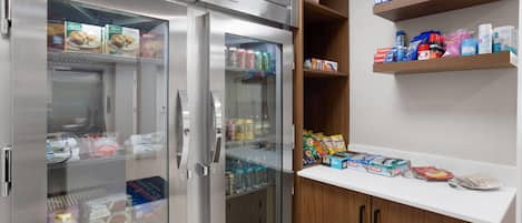 A kitchenette with a stainless steel refrigerator, a microwave, and a wooden cabinet with shelves holding various food items and beverages.