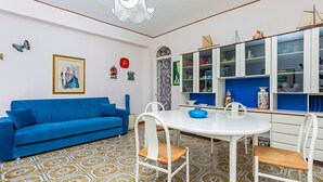 Quarto com sofá azul, cadeiras brancas, mesa branca, piso de azulejos estampados, luminária de teto branca e um armário azul com portas de vidro.