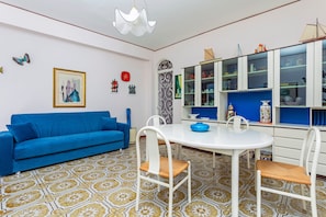 Une pièce avec un canapé bleu, des chaises blanches, une table blanche, des carreaux de sol à motifs, un luminaire blanc au plafond et un meuble bleu avec des portes en verre.