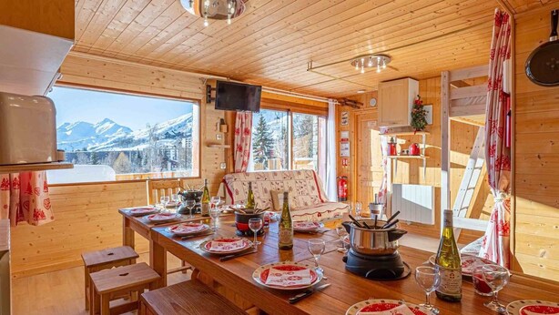 A wooden interior with a dining table set for four, a television, and a view of mountains. - Chalet 'Ground Floor 110 m² – Very Close to the Slopes' with Mountain View, Private Terrace & Wi-Fi (Fontcouverte-la-Toussuire)