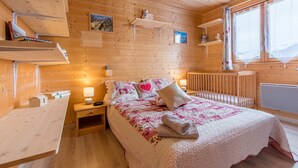 A wooden cabin bedroom with a bed, bedside tables, a nightstand, a crib, and a window with curtains. - Chalet 'Ground Floor 110 m² – Very Close to the Slopes' with Mountain View, Private Terrace & Wi-Fi (Fontcouverte-la-Toussuire)