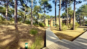 A wooden pathway leading to a building with multiple floors, surrounded by tall pine trees. - Cozy apartment between lake and ocean – Terrace & shared pool in Carcans-Maubuisson (Carcans)