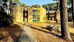A wooden pathway leading to a building with multiple balconies and a bicycle parked nearby. - Cozy apartment between lake and ocean – Terrace & shared pool in Carcans-Maubuisson (Carcans)