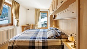 A bedroom with a bed, wooden headboard, a window with curtains, a radiator, and a desk with a lamp. - Apartment 'Genziana' with Mountain View, Private Terrace and Wi-Fi (Santa Caterina)