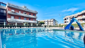 Una piscina con tobogán azul, sillas de descanso y un edificio con balcones.