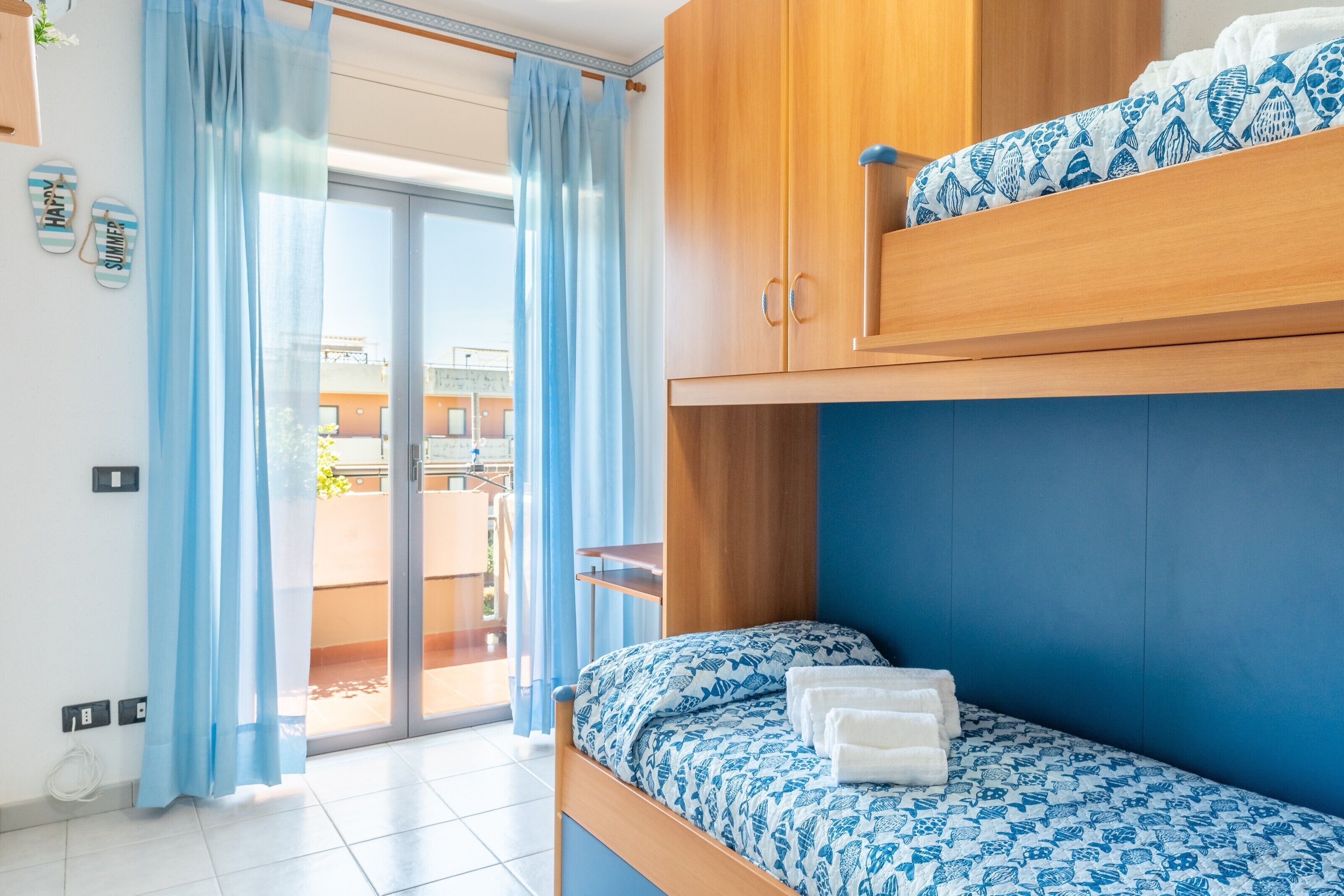 Una habitación con literas, una pared azul, un armario de madera y un ventanal corredero que da a un espacio exterior.