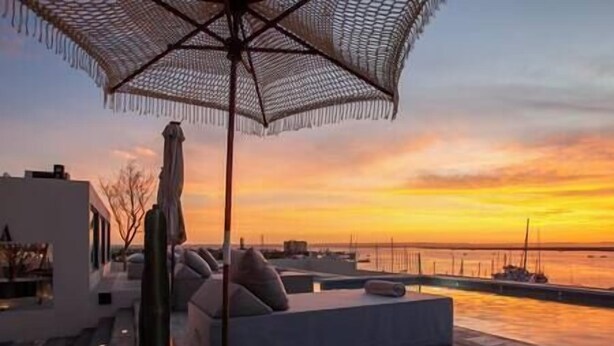 Een dakterras met uitzicht op de ondergaande zon over het water, met een loungehoek met kussens en een grote parasol.