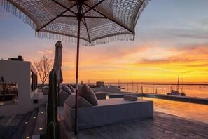 Eine Dachterrasse mit Blick auf den Sonnenuntergang über dem Wasser, ausgestattet mit einer Lounge-Ecke und einem großen Sonnenschirm.