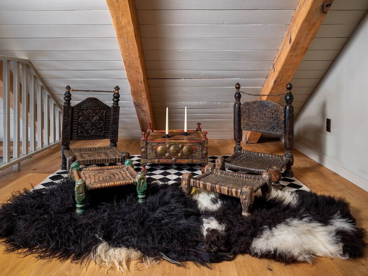 Ein rustikaler Raum mit hohen Decken und freiliegenden Balken, ausgestattet mit einem großen Bett mit Holz-Kopfteil, zwei Stühlen, einem kleinen Tisch und einem schwarz-weiß karierten Teppich.