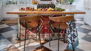 Een keuken met een centraal eiland, houten stoelen, een geruit vloertje en een raam met blinds.